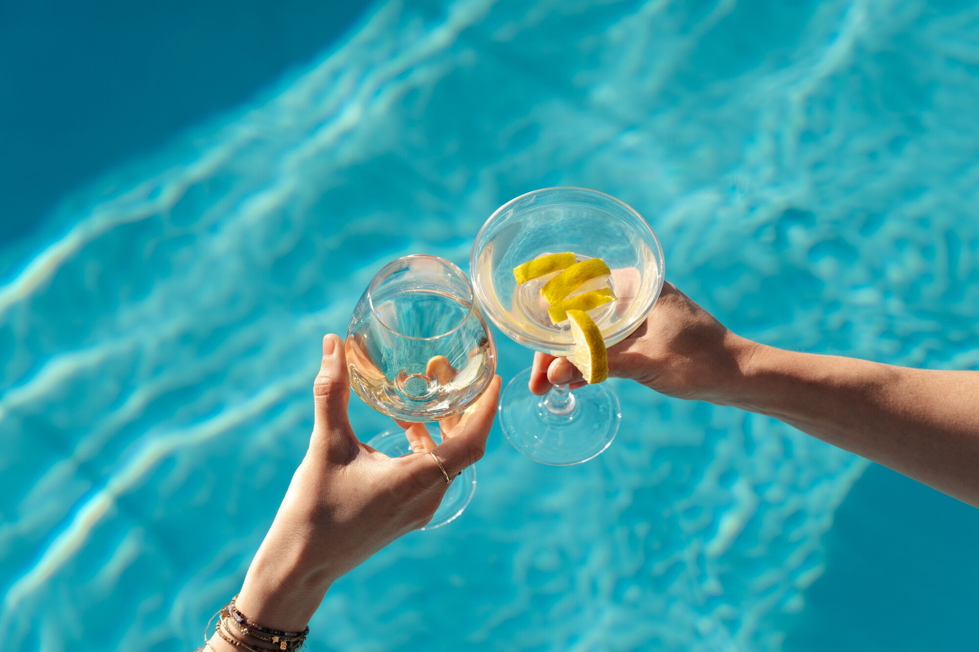 Deux mains tenant des verres transparents ornés d’une tranche de citron, sur fond d’une piscine bleue.