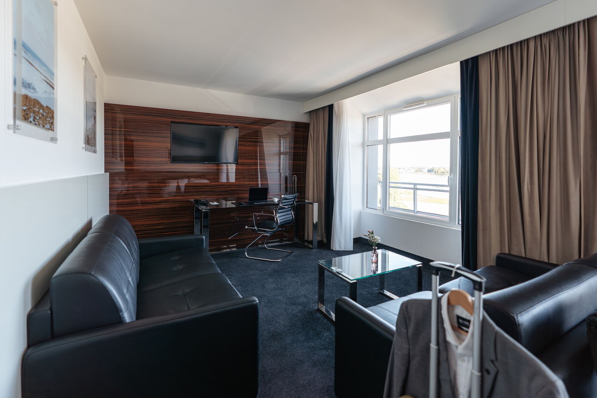 A modern hotel room with a dark leather sofa, a glass coffee table, a flat-screen TV mounted on a wooden wall, and a large window with curtains.
