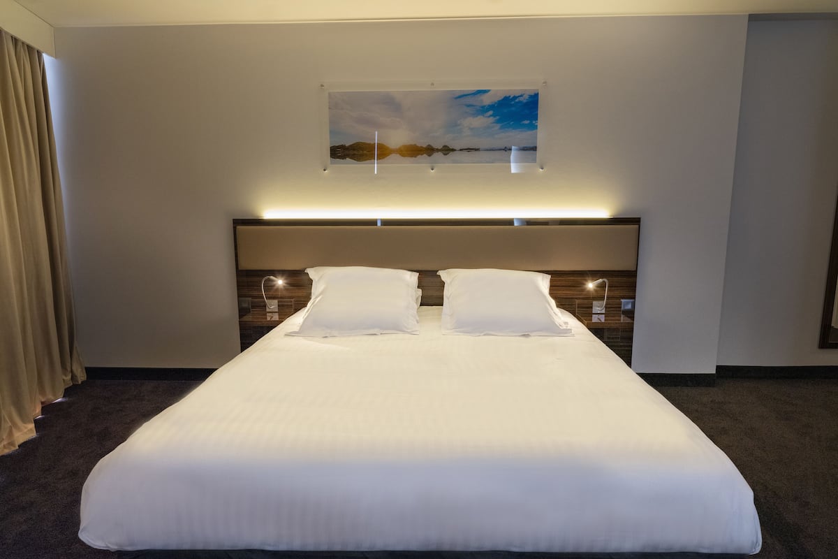 Quarto de hotel com uma cama grande, duas almofadas, cabeceira com iluminação embutida, um quadro sobre a cama e mesa de cabeceira com abajur.