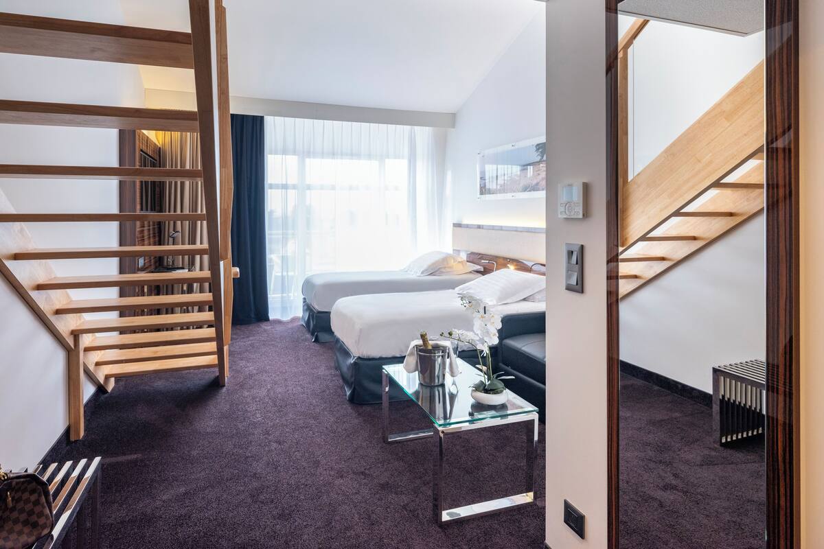 Quarto de hotel com escada de madeira, duas camas, uma mesa de centro de vidro e vista para o exterior.