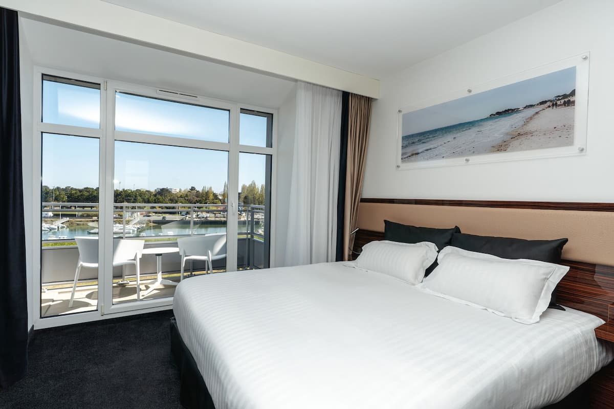 Quarto de hotel com uma cama grande, vista para a praia e varanda com cadeiras.