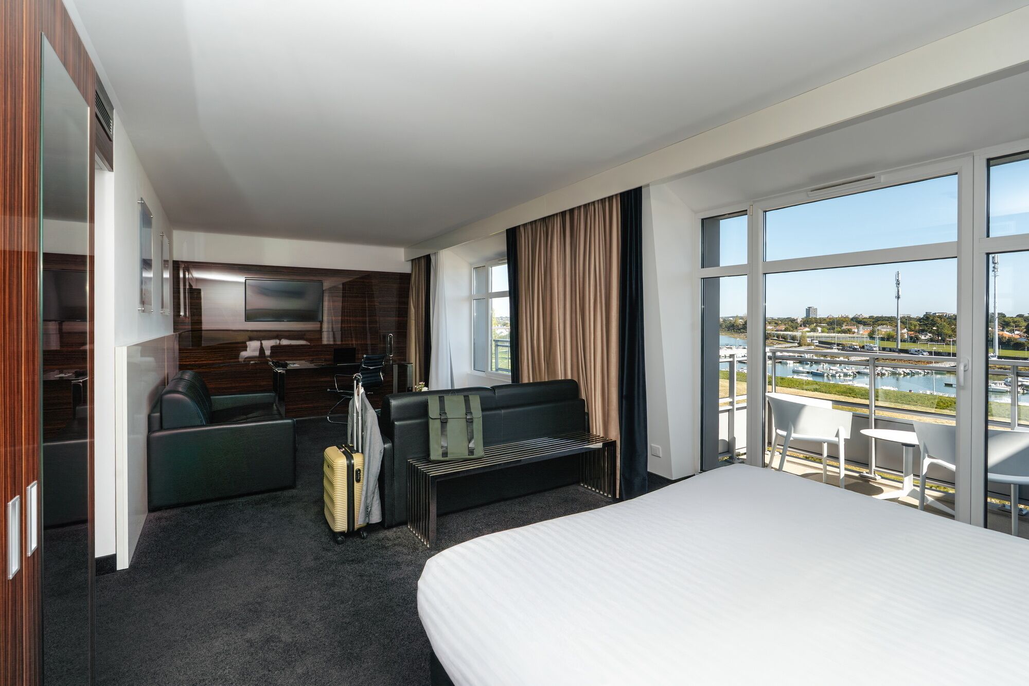 A hotel room with a bed, a seating area, a TV, a desk, and a large window with a view of a body of water and buildings.