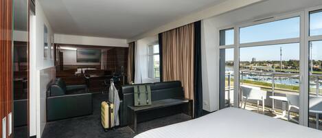A hotel room with a bed, a seating area, a TV, a desk, and a large window with a view of a body of water and buildings.