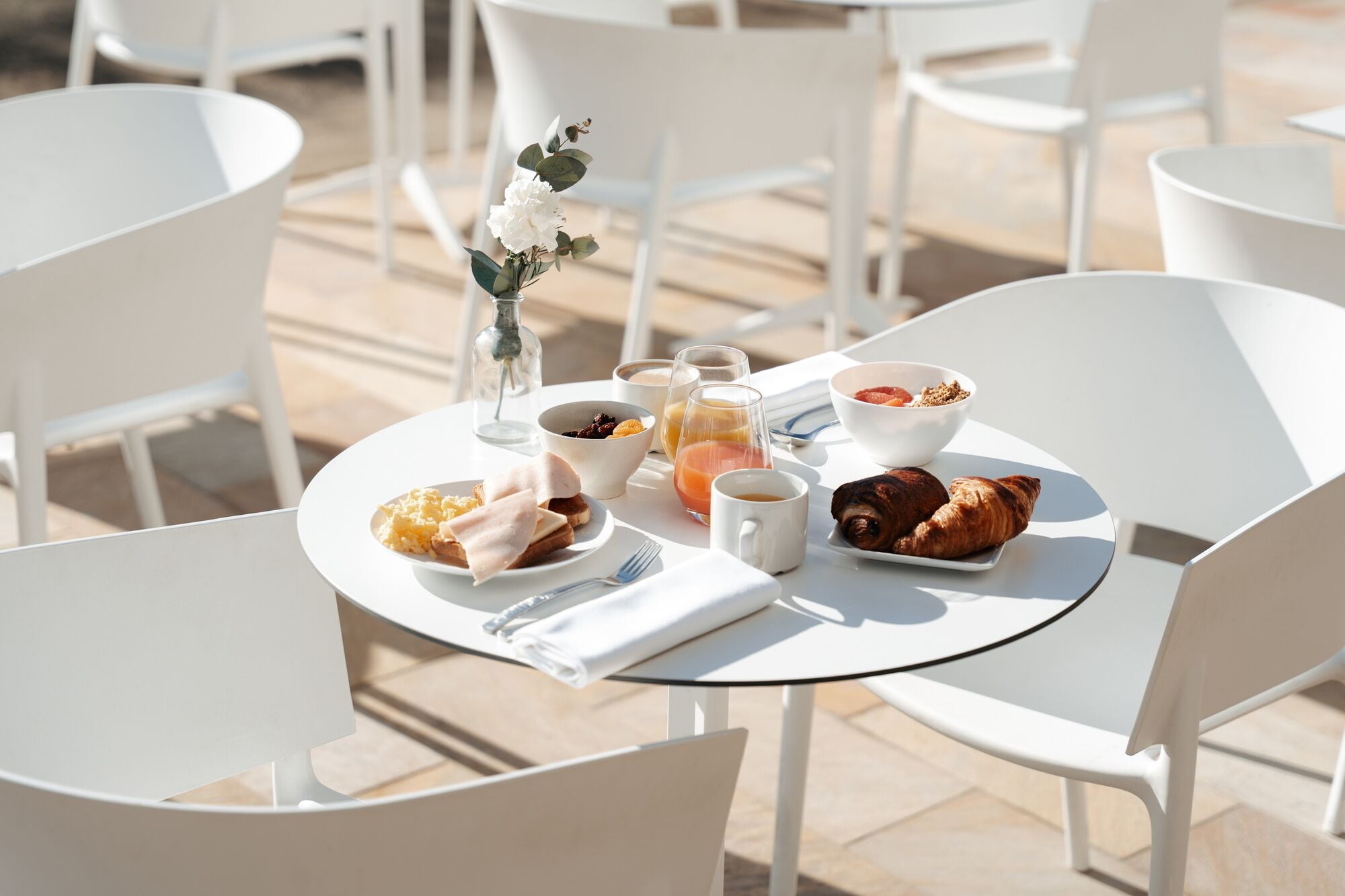 Une table ronde est dressée avec des aliments pour le petit-déjeuner, notamment des croissants, des toasts et des fruits. Des chaises blanches sont disposées autour de la table, et un vase rempli de fleurs y est posé.