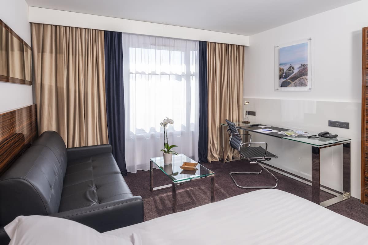 Quarto de hotel com uma cama, um sofá, uma mesa de escritório, uma cadeira, uma mesinha com uma planta e cortinas.