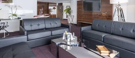A modern living room with a large flat-screen TV, two black leather sofas, a coffee table, and a small dining area with a table and chairs.