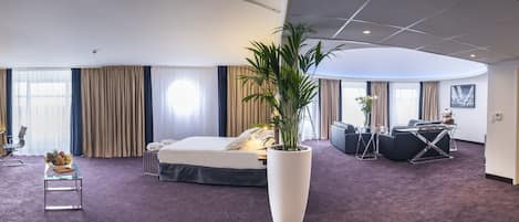 A modern hotel room with a bed, a desk, a chair, a large potted plant, and a view of the city.