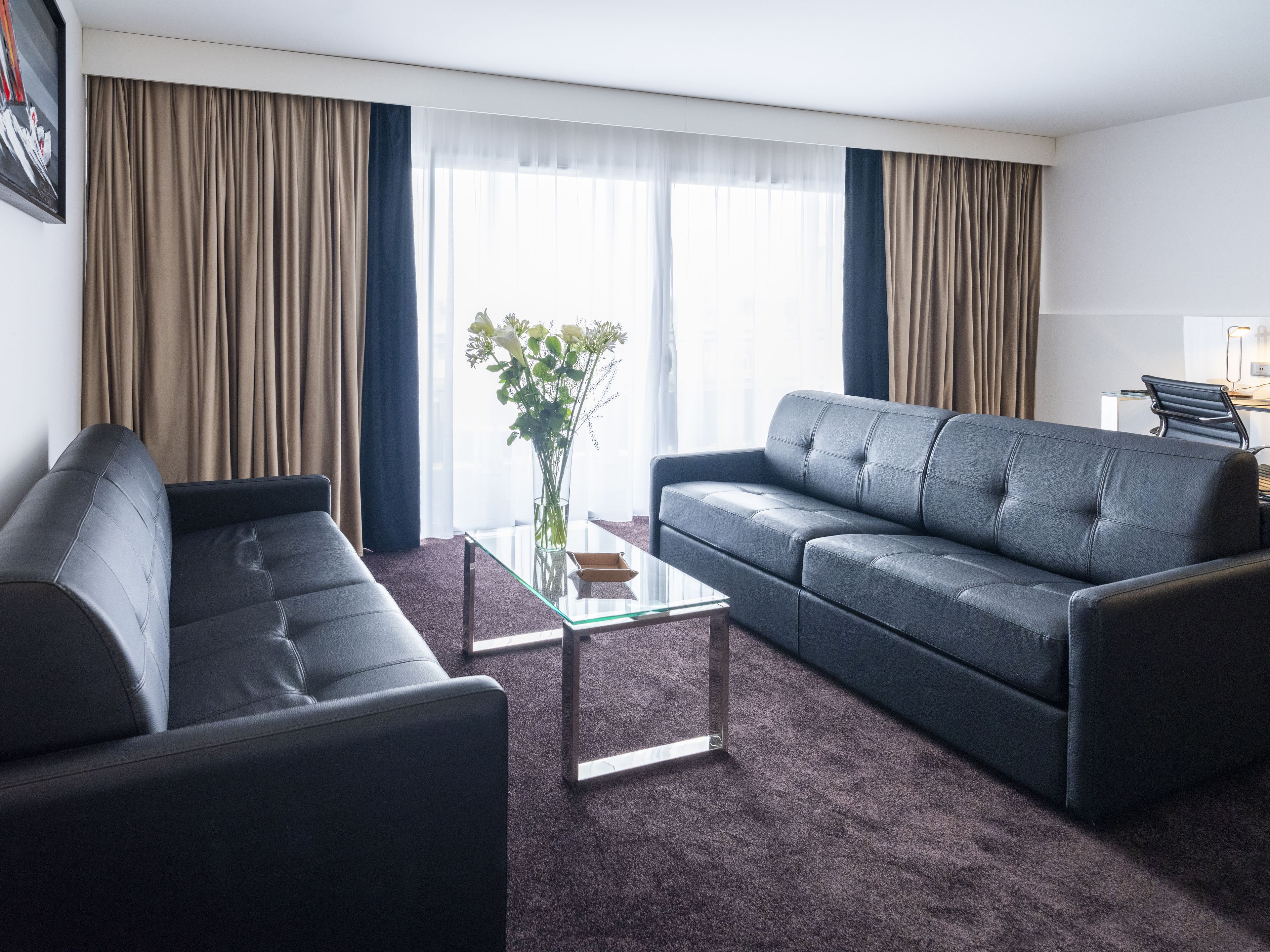 Een moderne woonkamer met een donker leren bank, een glazen salontafel en een bloemenvaas.