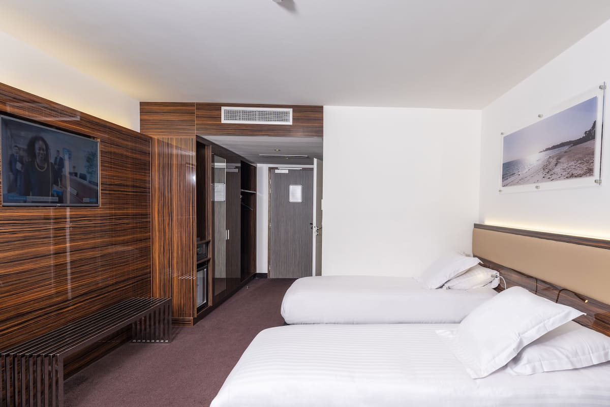 Quarto de hotel com duas camas, televisão, parede revestida de madeira, quadro emoldurado e porta para um banheiro.