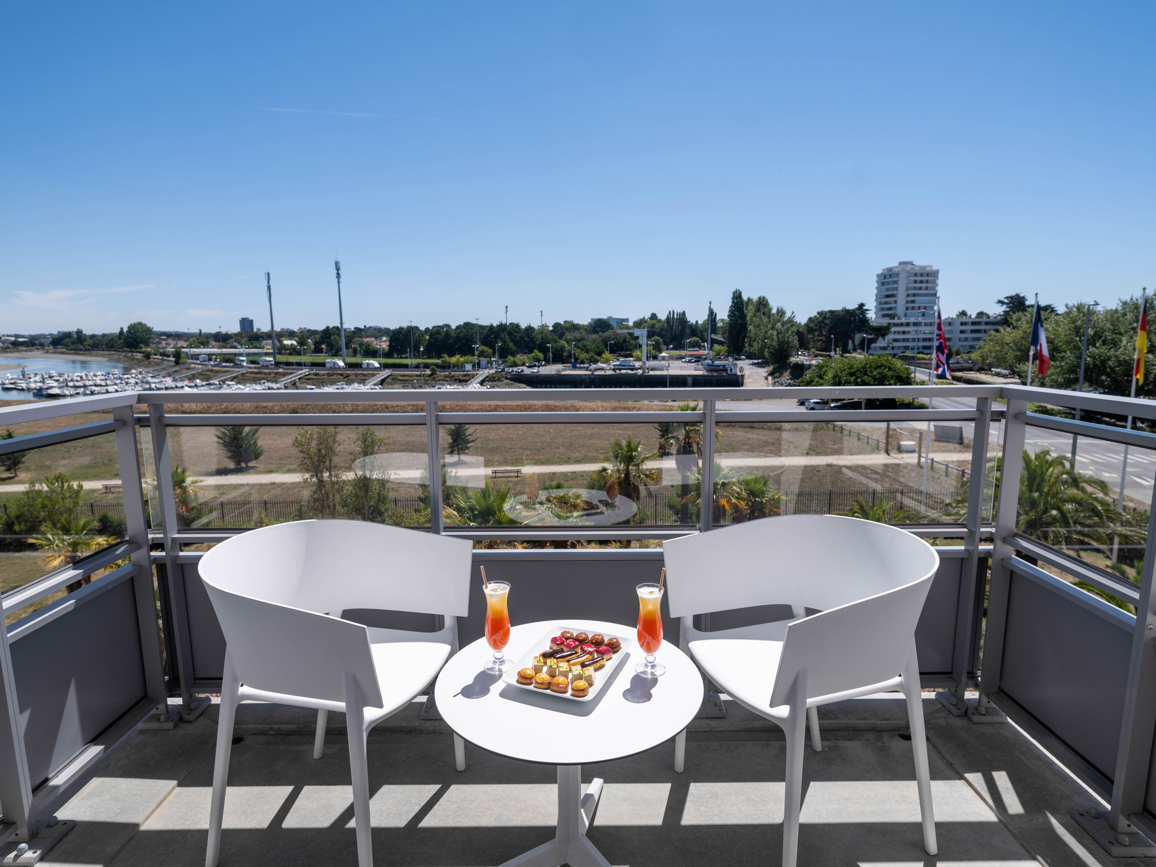 A balcony with two white chairs and a small table, overlooking a marina with boats and a cityscape in the background.