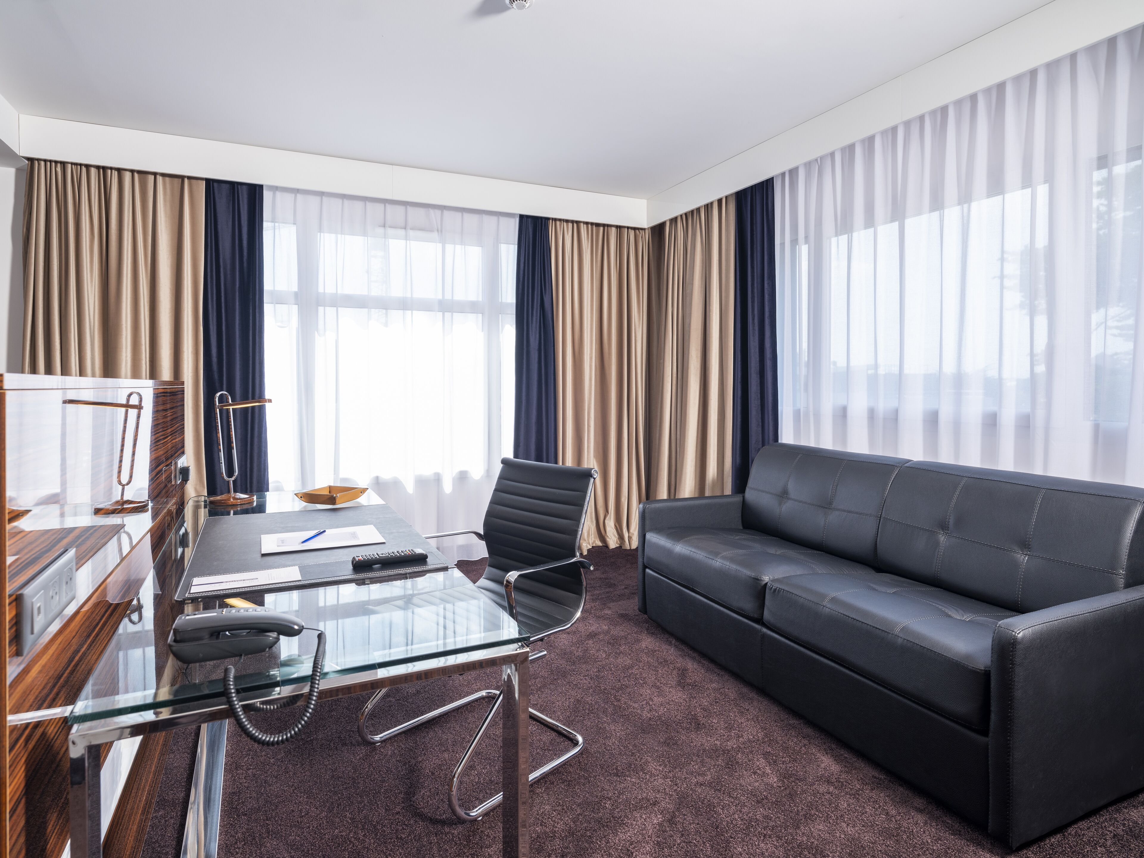 A modern hotel room with a glass table, a black leather sofa, and a large window with sheer curtains.