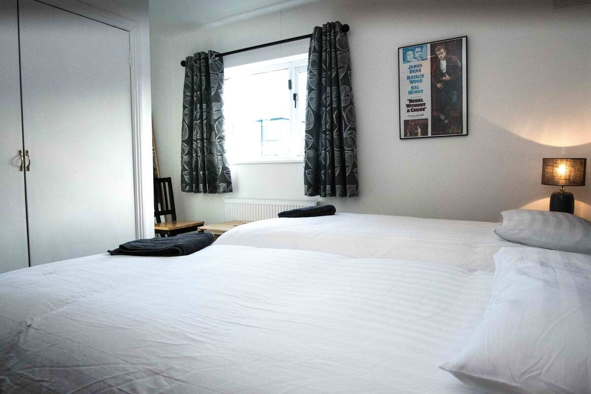 Un dormitorio con dos camas, una ventana con cortinas, un cartel en la pared y una lámpara en la mesita de noche.