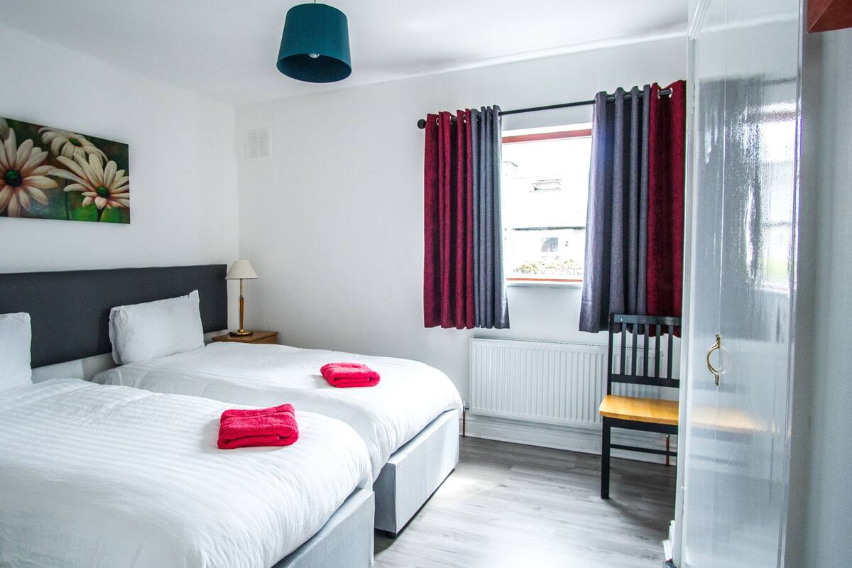 Habitación de hotel con dos camas, un cuadro de flores en la pared, una ventana con cortinas y una silla junto a la ventana.