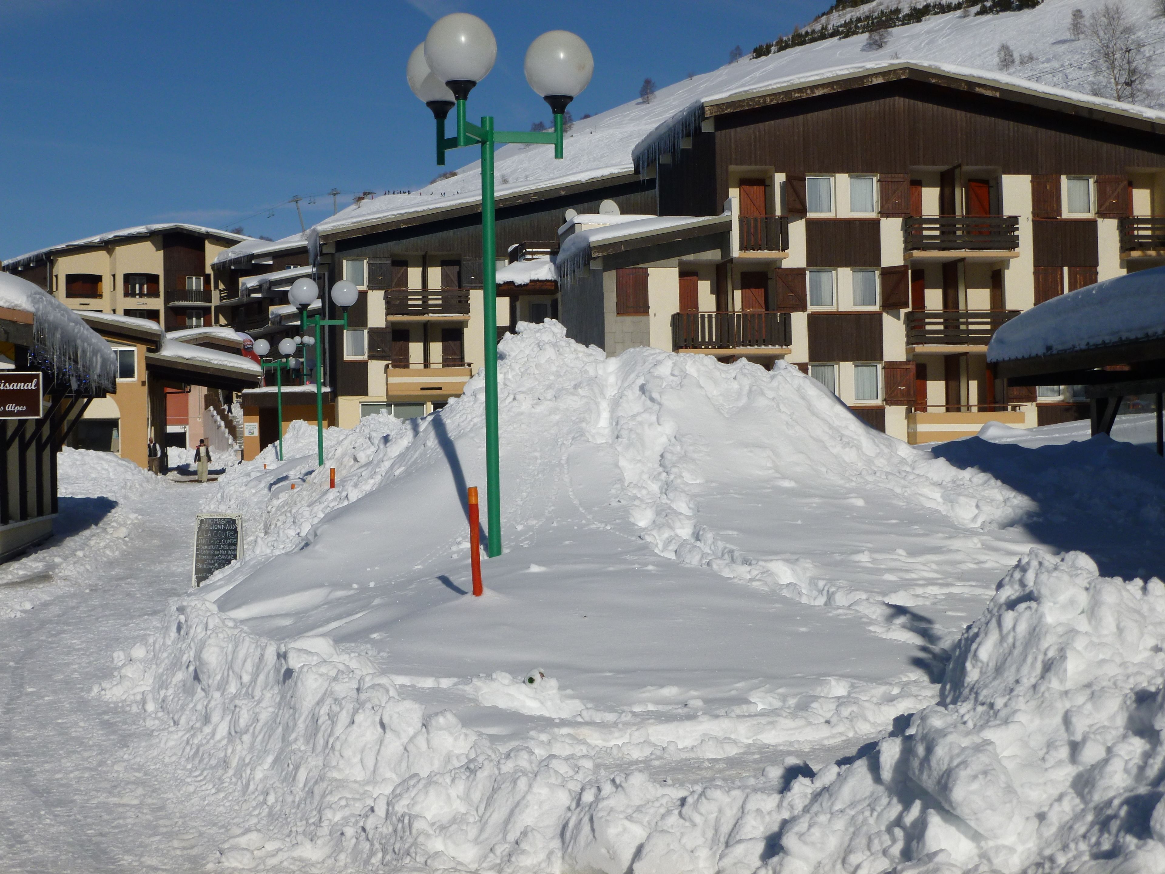 A snow-covered outdoor area with a pile of snow, a green street lamp, and a building with balconies in the background.