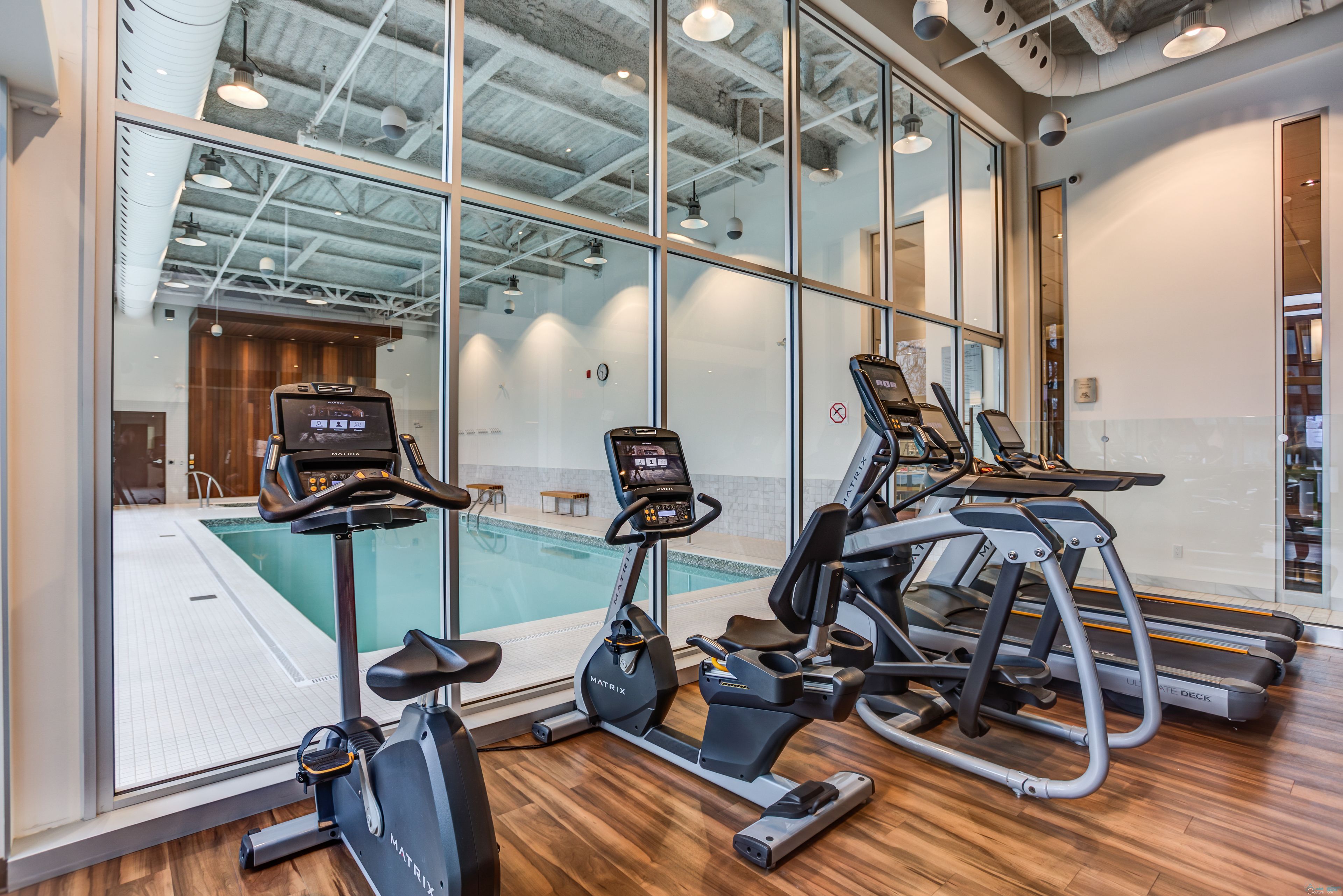 A gym with treadmills, an elliptical, and a pool visible through large windows.
