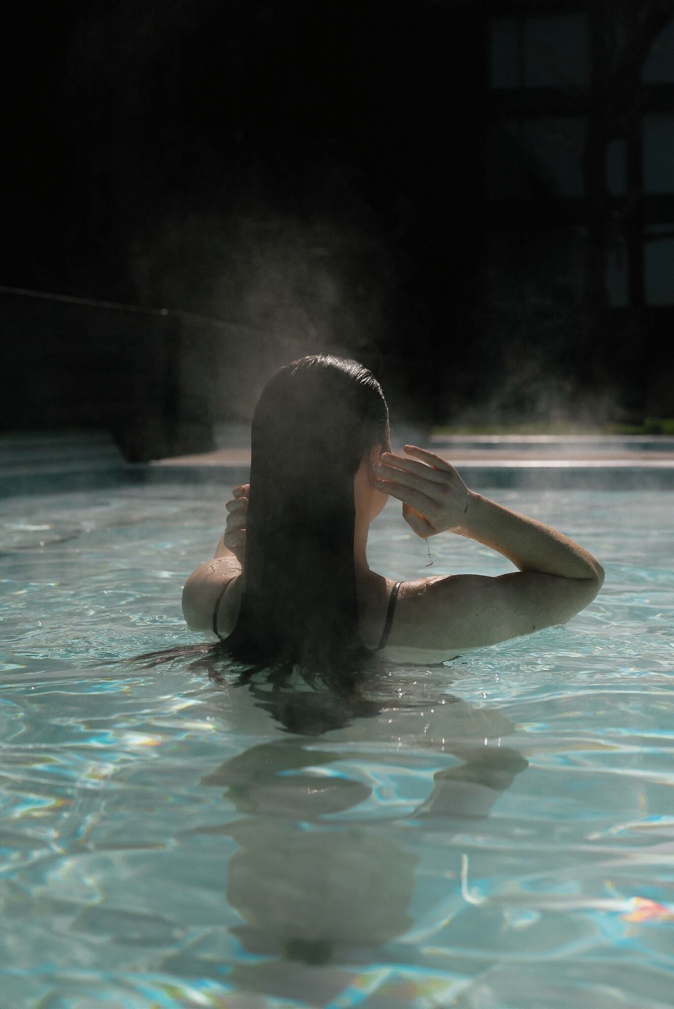 A person in a swimming pool with clear water, steam rising from the pool, and a building in the background.