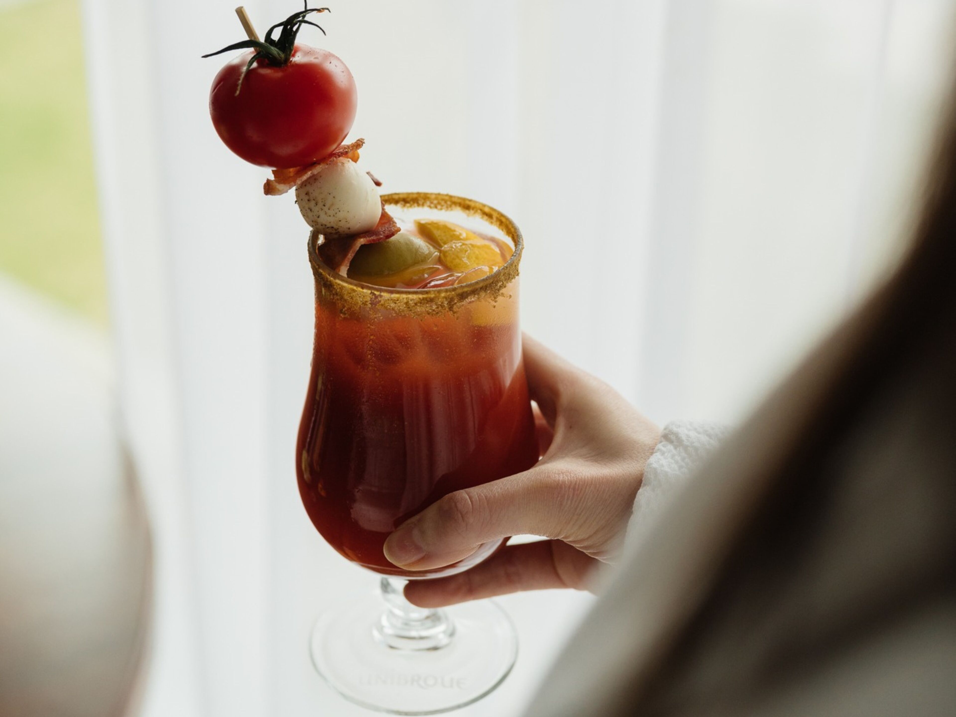 A person holding a Bloody Mary cocktail with garnishes.