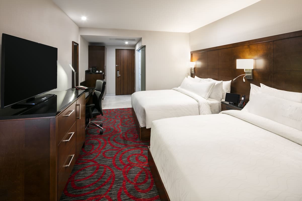 Ein Hotelzimmer mit Fernseher, Schreibtisch mit Schubladen, zwei Betten mit weißer Bettwäsche, einem roten und schwarzen Teppich mit Muster sowie einem Holz-Kopfteil.