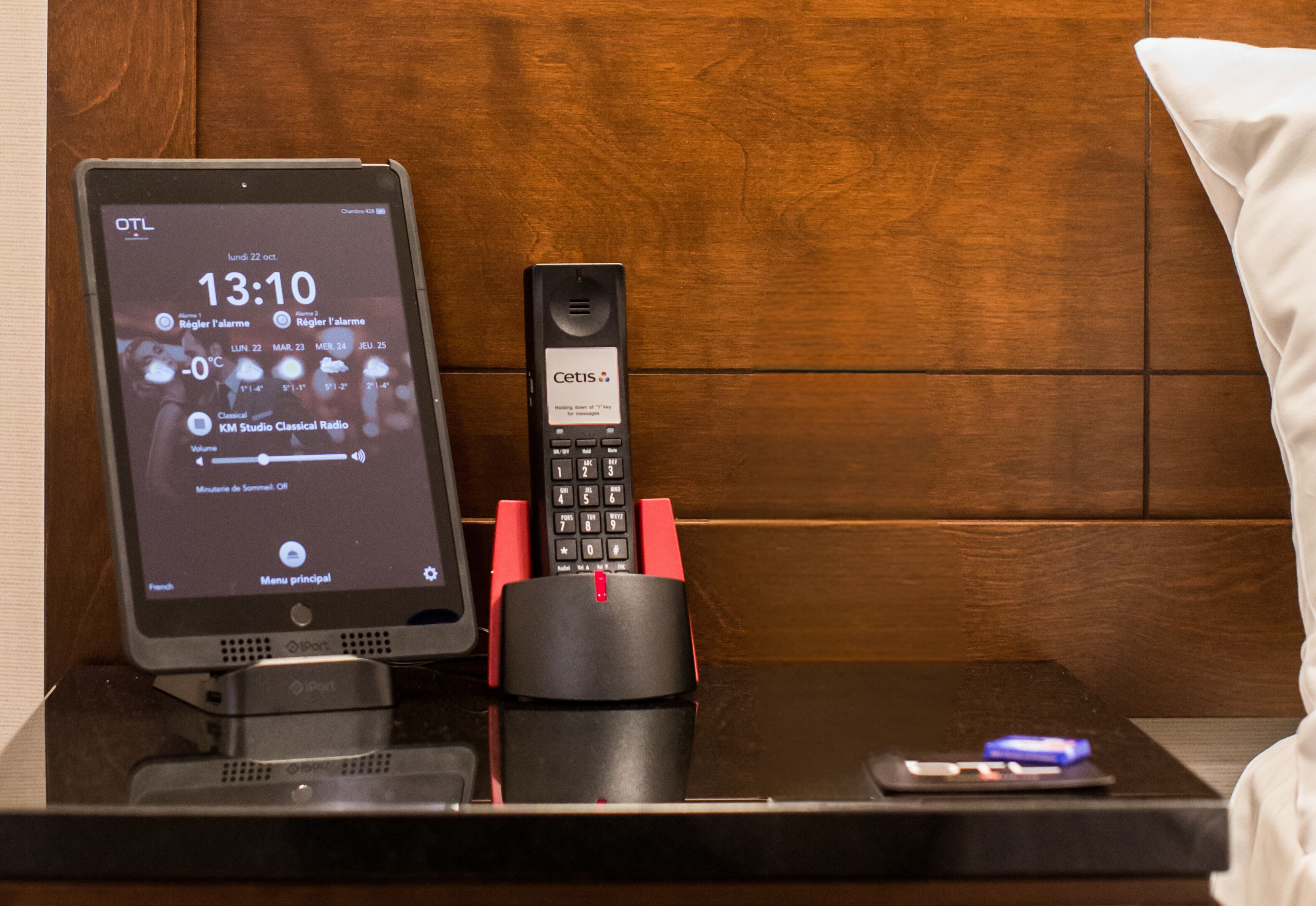 Habitación de hotel con cabecera de madera, mesita de noche con una tableta que muestra la hora y el clima, teléfono fijo y control remoto.
