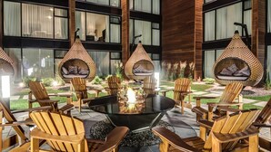 Un espacio exterior moderno con una zona de estar, una mesa redonda de vidrio rodeada de sillas de madera y una chimenea central.