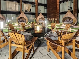 A modern outdoor seating area with a round glass table surrounded by wooden chairs and a central fire feature.