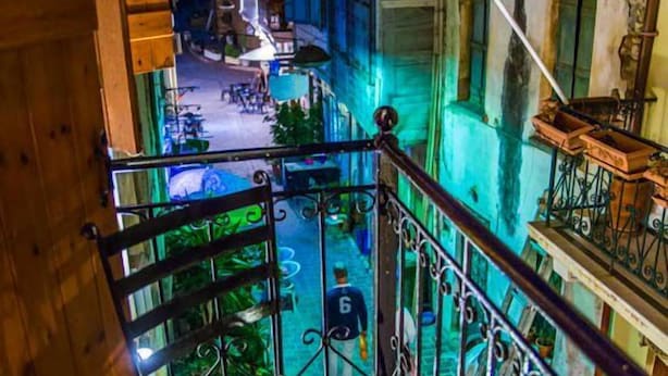 A narrow alleyway with buildings on either side, a balcony with a metal railing, and a street with outdoor seating visible in the distance. - Villa Milli (Chania)