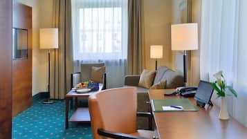 A hotel room with a desk, two chairs, a sofa, a lamp, a phone, a vase with flowers, and a window with curtains.