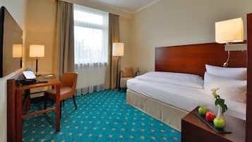 A hotel room with a bed, desk, chair, and a small table with apples and a vase.