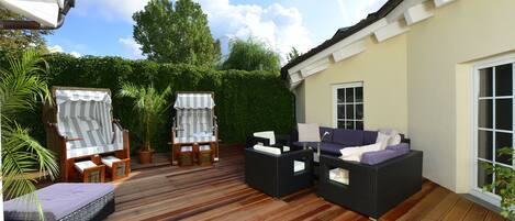 A deck with outdoor furniture, including a sofa and chairs, surrounded by greenery and a building.