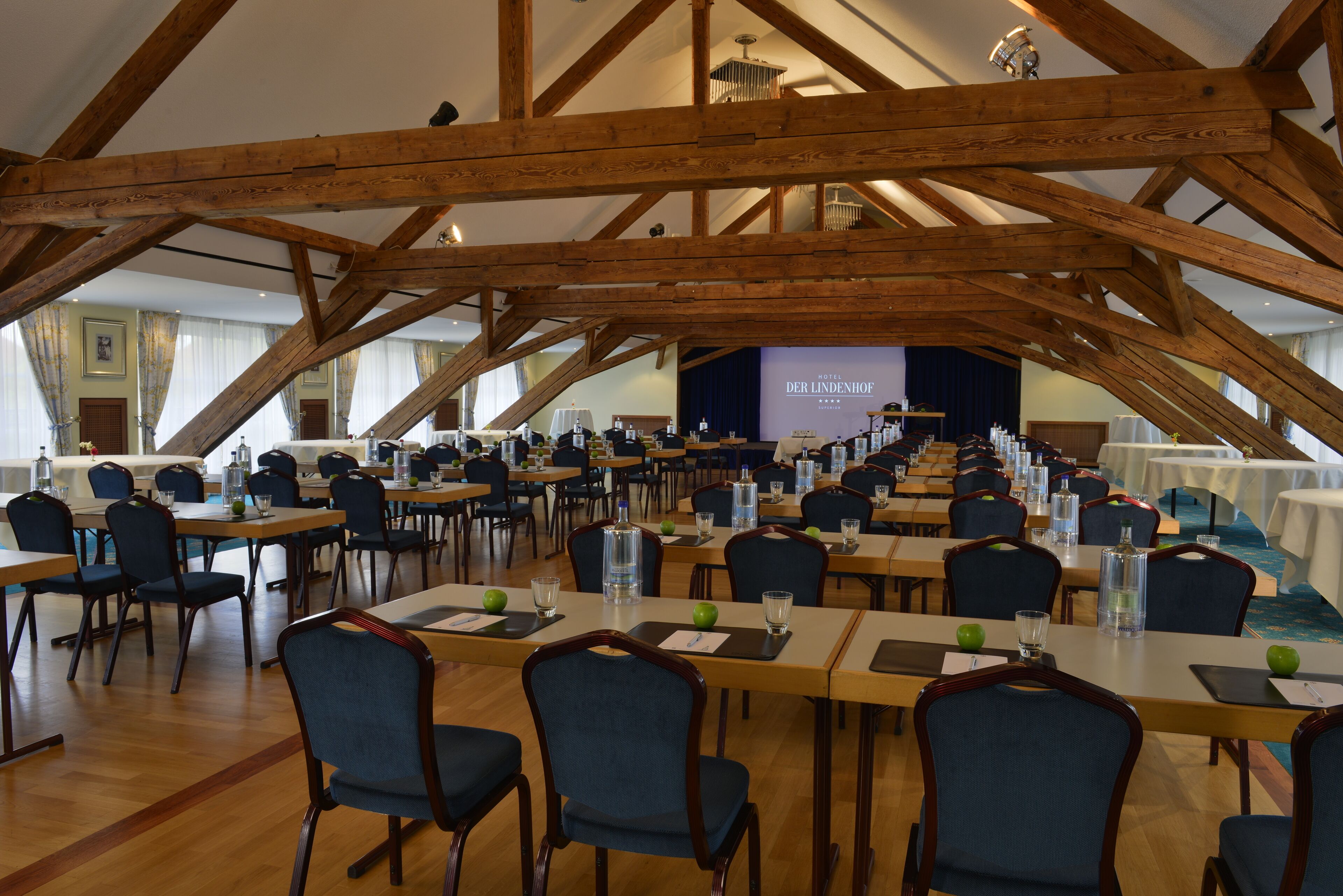 Een ruime hal met houten balken, tafels met stoelen en een podium met een projectiescherm.