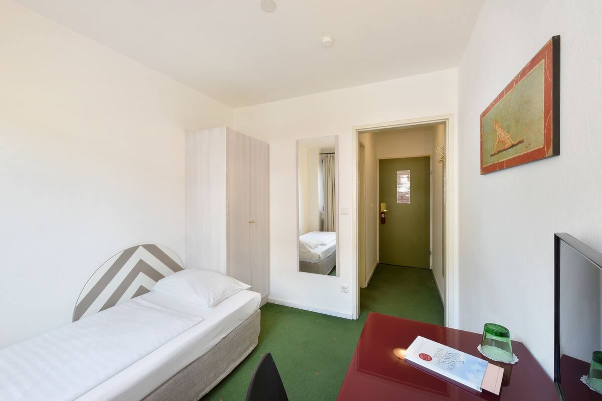 A hotel room with a bed, a desk with a lamp, a mirror, and a door leading to another room.