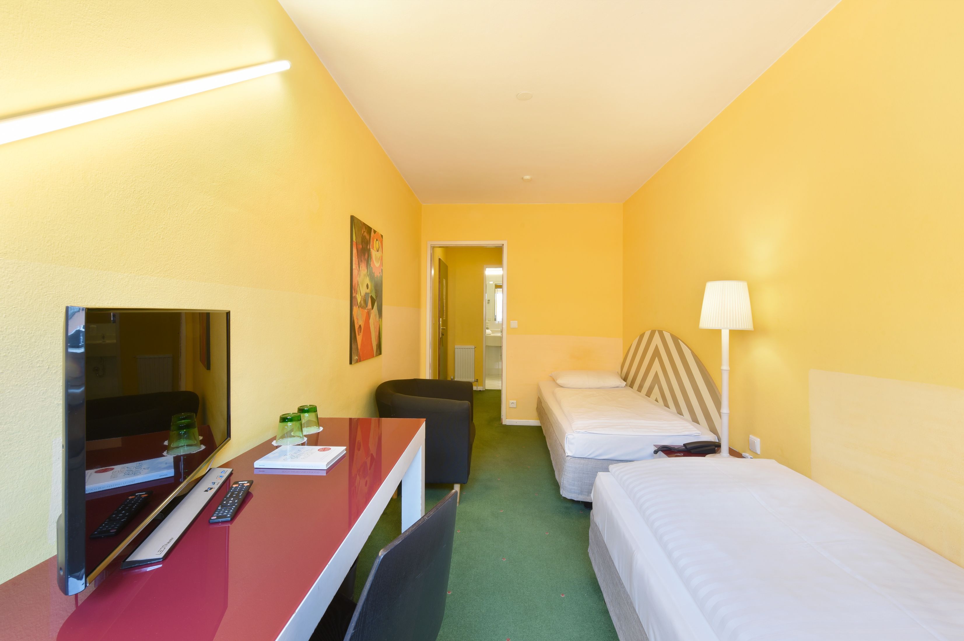 Habitación de hotel con escritorio, dos camas, televisión, lámpara y un cuadro en la pared.