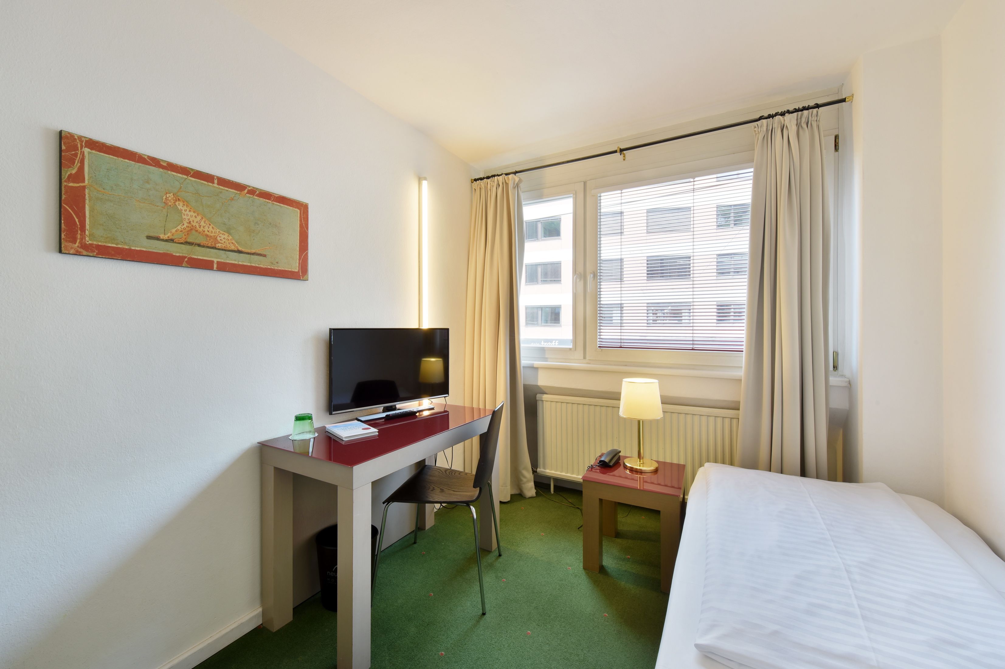 Ein Hotelzimmer mit Bett, Schreibtisch, Stuhl, Fernseher, Lampe und einem Bild an der Wand.