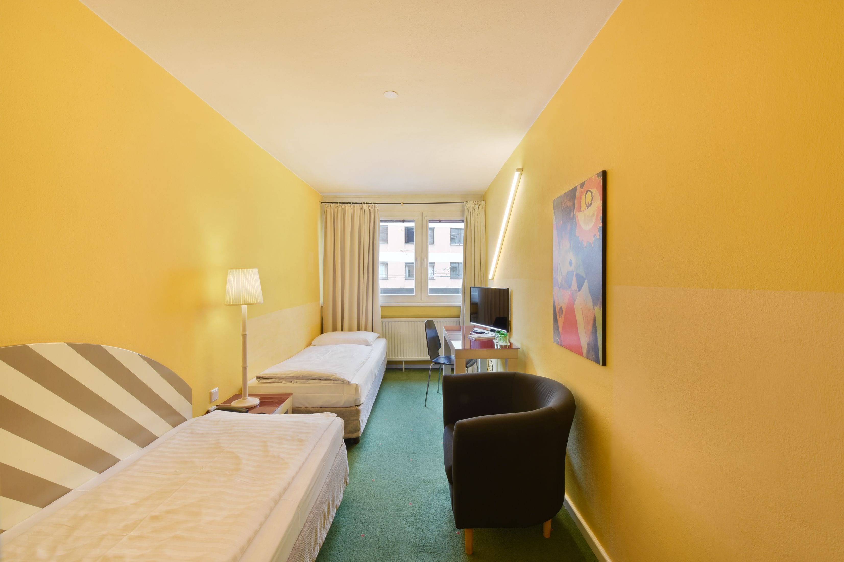 Habitación de hotel con dos camas, un escritorio, una silla, una lámpara, una ventana con cortinas y un cuadro en la pared.