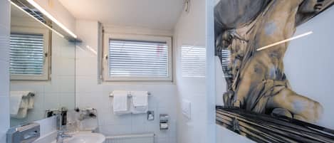 A modern bathroom with a large mirror, white tiles, a sink, and a window with blinds.