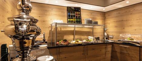 A modern kitchen with a stainless steel coffee machine, a glass display case with food items, wooden cabinetry, and a ceiling with recessed lighting.