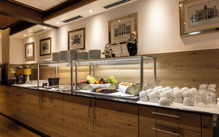 A buffet area with a glass display case containing fruits, a selection of cheeses, and a variety of breads.