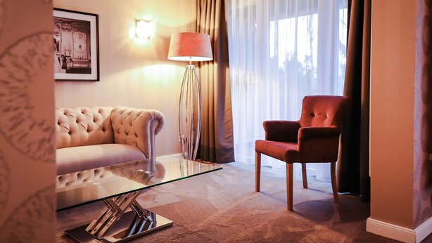 Une chambre d’hôtel avec un canapé capitonné, un fauteuil rouge, une table basse en verre, une lampe sur pied et un tableau encadré au mur.