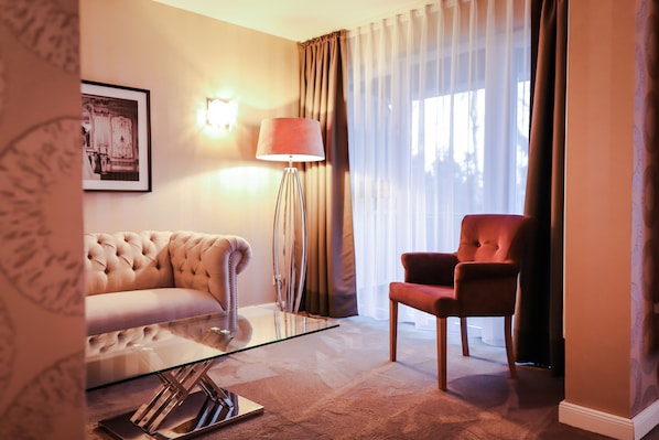 Habitación de hotel con un sofá capitonado, un sillón rojo, una mesa de centro de vidrio, una lámpara de pie y un cuadro en la pared.