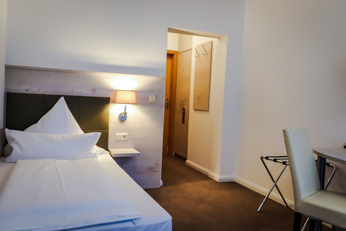 Habitación de hotel con una cama bien hecha, una lámpara de noche, una silla y una puerta que da a otra habitación.