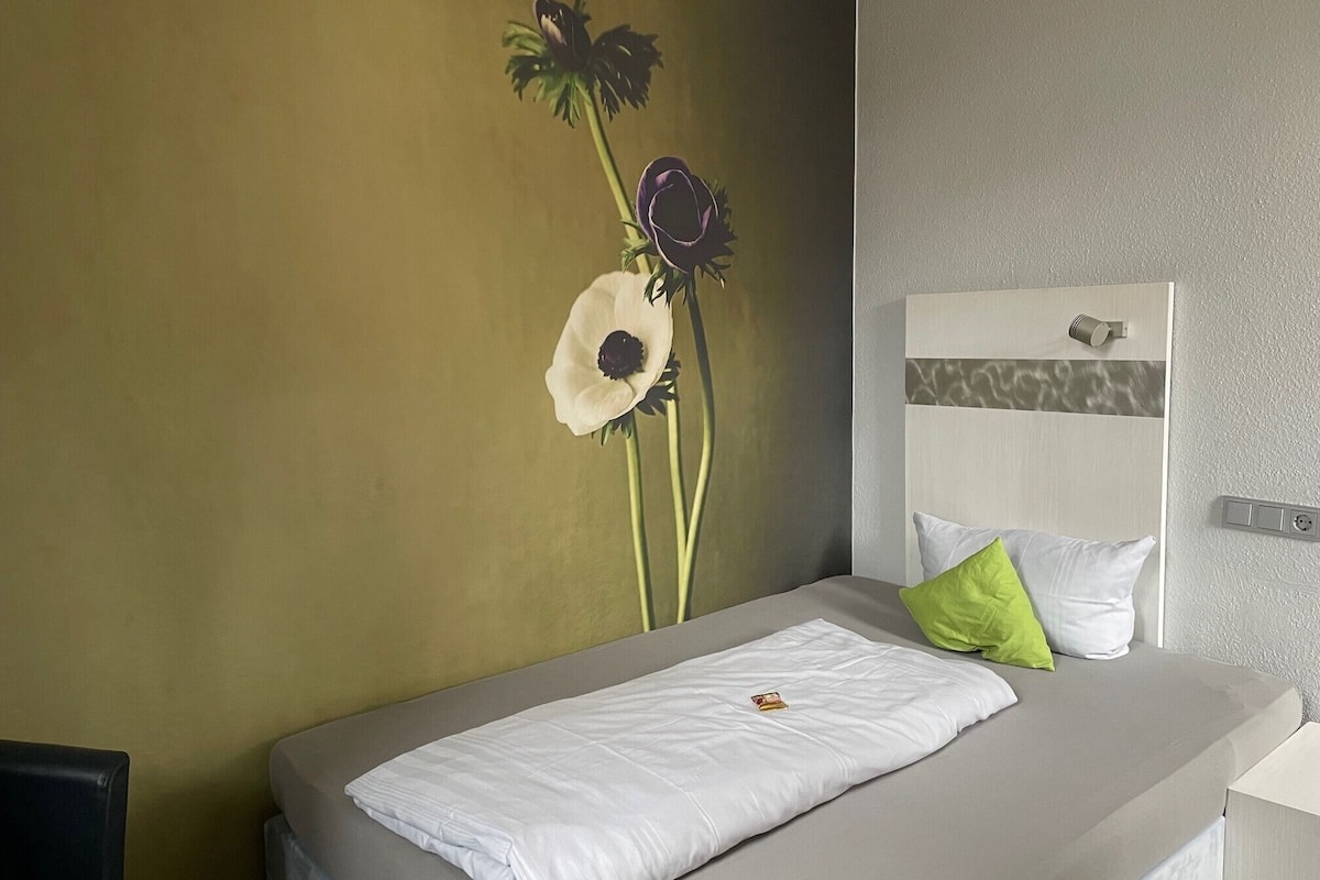 Una cama individual con colchón y almohadas blancos, una mesita de noche pequeña y una lámpara fijada a la pared.