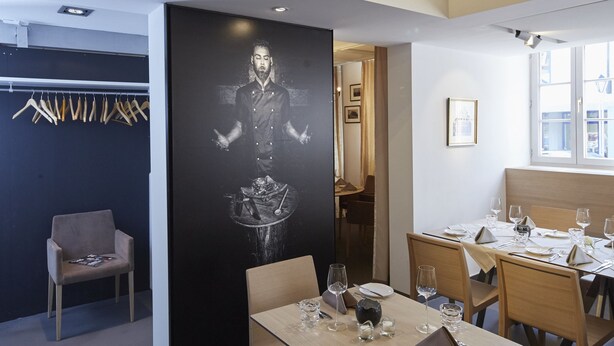A modern dining area with a high ceiling, wooden beams, and a black wall with a portrait. Tables are set with wine glasses and plates. - Hotel & Restaurant Badischer Hof (Bühl)