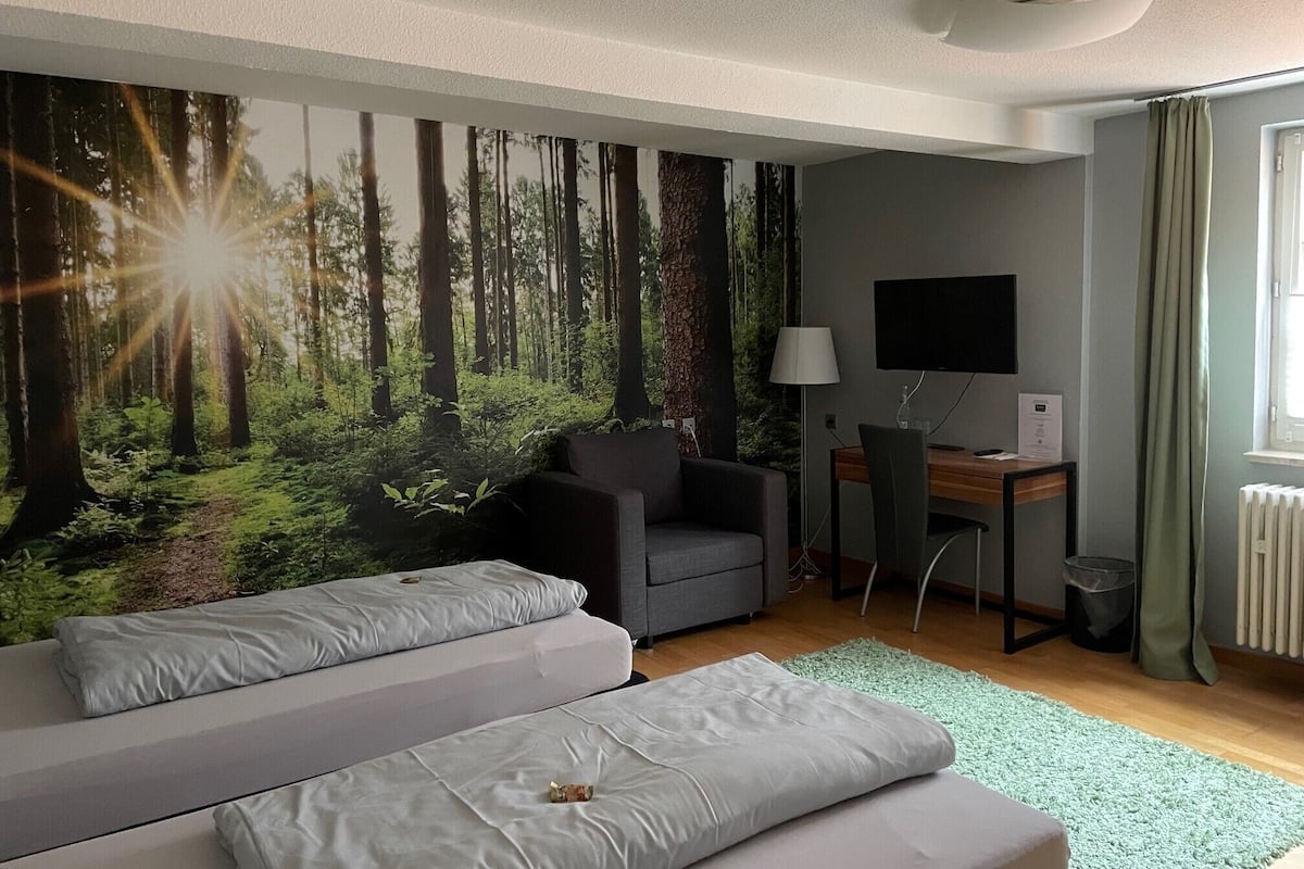 Un dormitorio con un gran mural de un bosque, una cama, un escritorio con una computadora y un televisor.