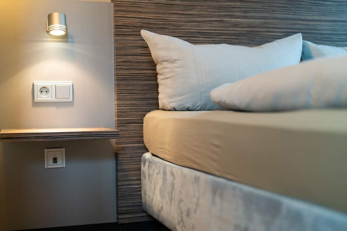 Un dormitorio ordenado con cama, almohadas, una mesita de noche y una lámpara de pared.