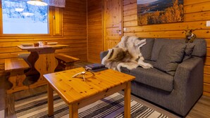 A cozy cabin interior with a wooden interior, a sofa, a table with a lamp, and a window with a view of trees and a bridge.