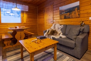 Interior acogedor de una cabaña con detalles de madera, un sofá, una mesa con lámpara y una ventana que da a vista de árboles y un puente.