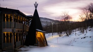 A lodge with a wooden exterior, a staircase leading to a teepee-like structure, and a snowy landscape with a mountain in the background.