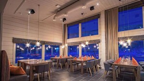 A modern dining area with multiple tables and chairs, large windows with a view, and pendant lighting.