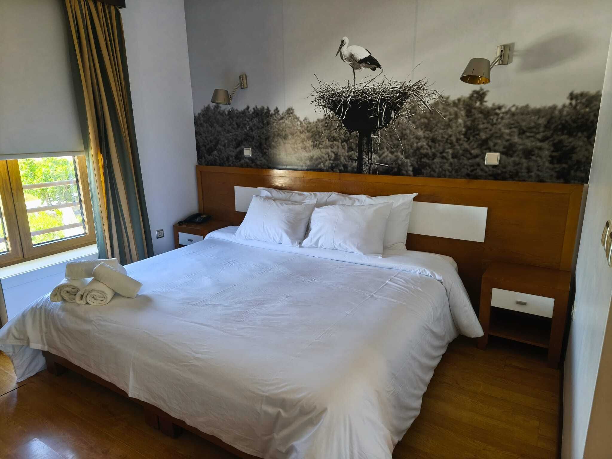 Habitación de hotel con una cama grande, una mesita de noche, una lámpara y una obra de arte en la pared con la imagen de un pájaro.