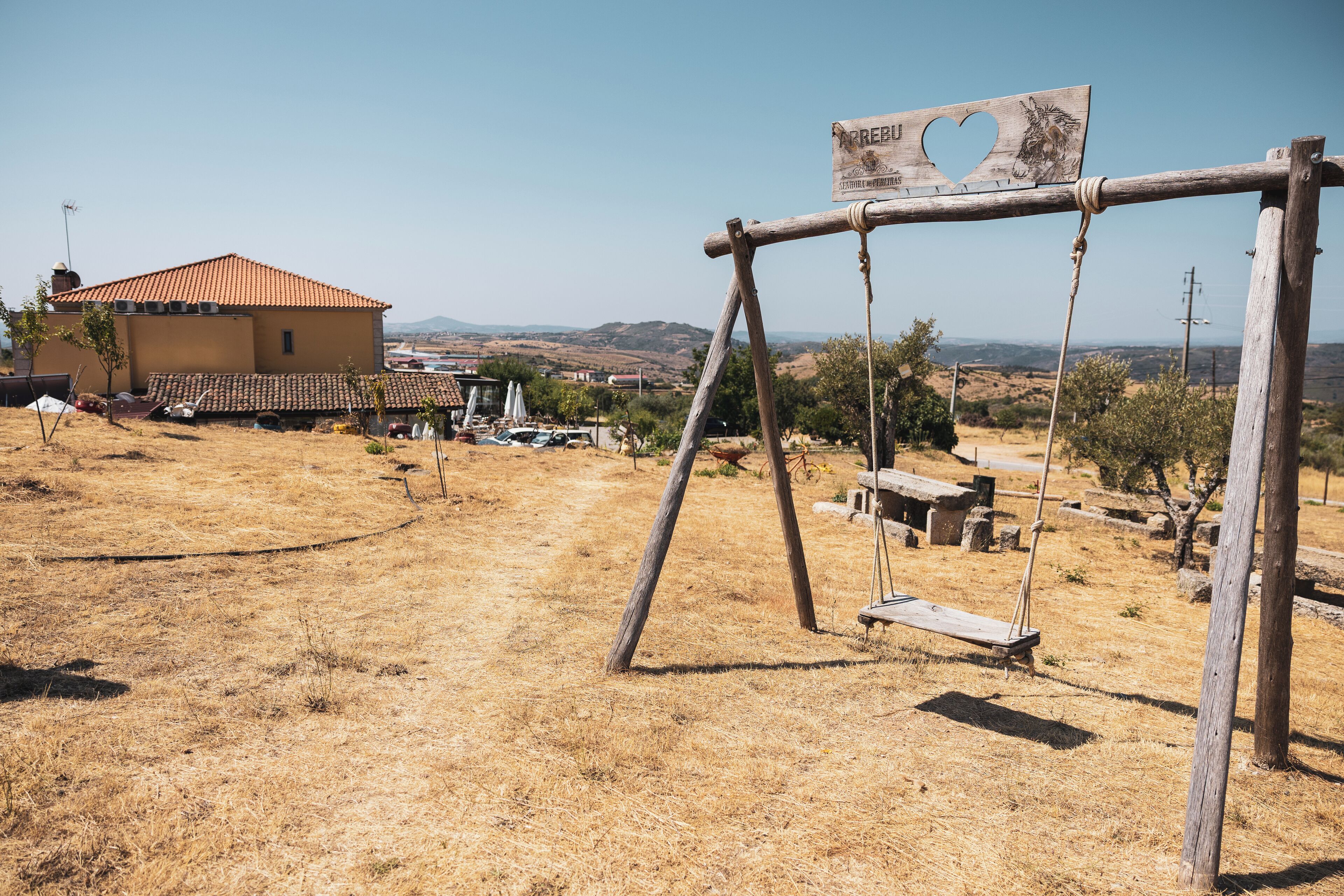 A rustic wooden swing set in a dry, open field with a house in the background.